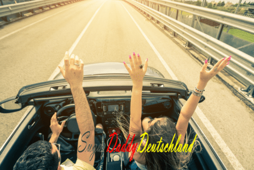 Hombre y mujer levantan los brazos en un descapotable y se divierten.